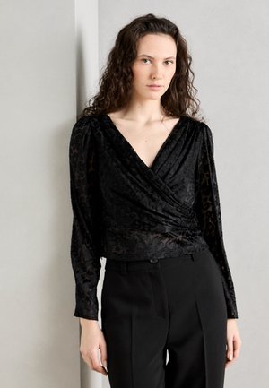 Black wrap blouse with sheer, textured fabric featuring a floral pattern. Long sleeves and a V-neckline, paired with tailored black pants.