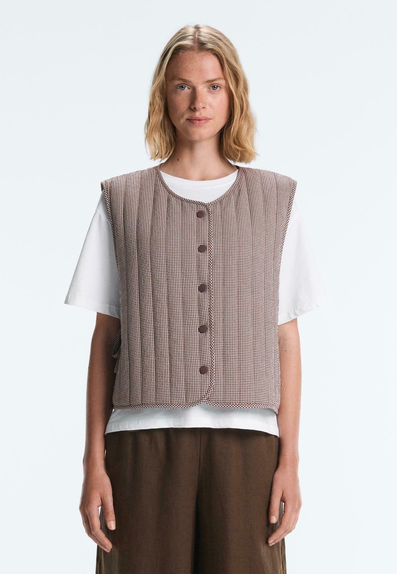 Femme portant un gilet sans manches marron à carreaux boutonné par-dessus une chemise blanche à manches courtes et un pantalon marron, debout devant un fond uni.