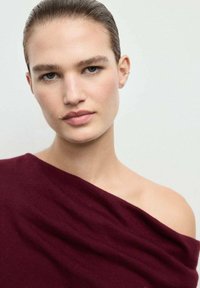 Crimson knit top with one shoulder exposed, featuring soft texture and subtle draping. Model has slicked-back hair and minimal makeup.