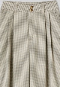 Pantalon beige à carreaux avec une taille haute, doté de deux boutons marron et de plis nets, fabriqué en tissu texturé.