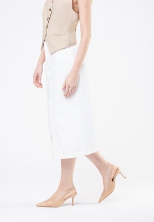 Gilet beige senza maniche con bottoni, abbinato a una gonna midi bianca e décolleté nudi in vernice con punta affusolata e tacco sottile.