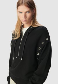 Felpa nera con cappuccio realizzata in tessuto morbido, con zip frontale, cappuccio e occhielli decorativi in metallo lungo le spalle. Presenta un cordino con punte in metallo.