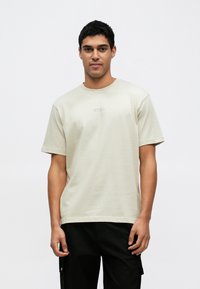 Trumpomis rankovėmis, smėlin�ės spalvos medvilniniai marškinėliai su apvalia iškirpte ir subtiliu logotipo motyvu ant krūtinės. Dėvimi su juodomis kelnėmis.