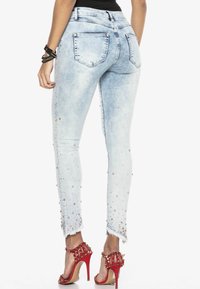 Jasnoniebieskie dżinsy o dopasowanym kroju z ozdobnym wykończeniem dołu, ozdobione metalowymi ćwiekami. Surowy brzeg na kostkach. Noszone z czerwonymi szpilkami.