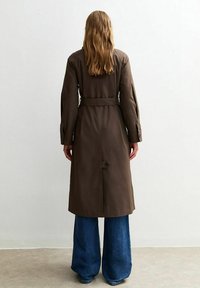 Brązowy płaszcz trench z paskiem w talii, długimi rękawami i guzikami, w połączeniu z szerokimi niebieskimi dżinsami. Gładka tekstura i zorganizowany design.