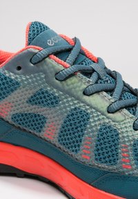 Teal en koraalkleurige sportschoen met mesh materiaal, textuuraccenten en een dikke zool met een looppatroon voor grip.