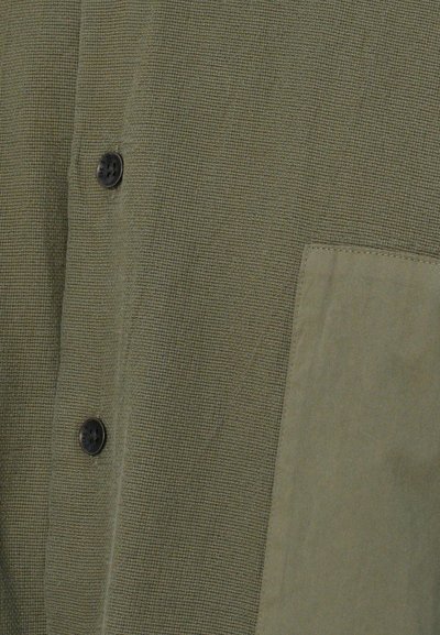Olivgrüne Button-Up-Hemd aus strukturiertem Stoff, mit zwei schwarzen Knöpfen und einer Leistentasche aus glattem Material auf der linken Seite.