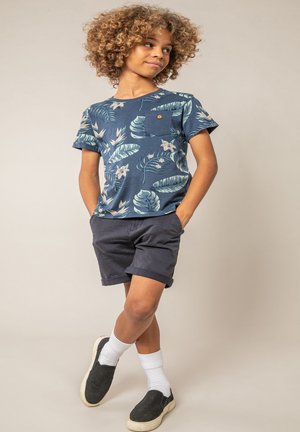 Enfant aux cheveux bouclés portant une chemise bleue à motifs floraux, un short foncé, des chaussettes blanches et des chaussures noires sans lacets, debout les mains dans les poches.