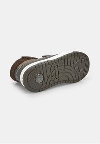 Pantof cu piele de nubuc maro, cu talpă din cauciuc; are curele Velcro și o talpă intermediară albă, evidențiată de un șanț texturat pentru aderență.