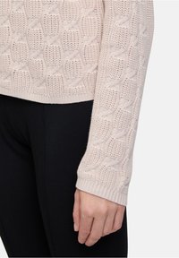 Hellbeige gestrickter Pullover mit strukturiertem Zopfmuster, gerippten Bündchen und lockerer Passform, kombiniert mit schwarzen Leggings.