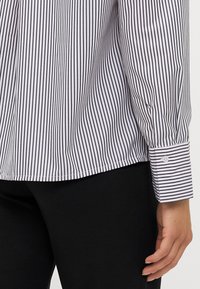 White and navy striped shirt featuring long sleeves with buttoned cuffs, smooth fabric, and a relaxed fit, paired with black pants.