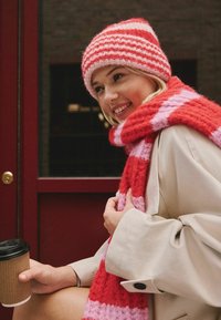 Red and pink striped knit hat and matching scarf, worn with a light beige coat. Holding a disposable coffee cup in a brown sleeve.