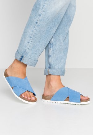 Blue suede cross-strap sandals with a cork footbed and white rubber outsole, worn with rolled-up light denim jeans.