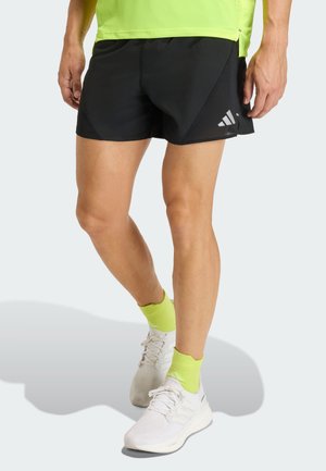 Man wearing black Adidas running shorts, white Adidas sneakers, and neon yellow socks, standing on a light gray surface.