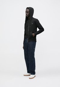 Sudadera con capucha negra con cremallera y un logo triangular, combinada con una camiseta negra y vaqueros azul oscuro. Zapatillas blancas con detalles en marrón completan el conjunto.