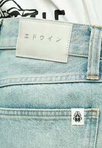 Φωτεινό μπλε τζιν από denim με λευκή δερμάτινη ετικέτα που φέρει ιαπωνικό κείμενο και μια μικρή ετικέτα με μαύρο λογότυπο σκαθαριού.