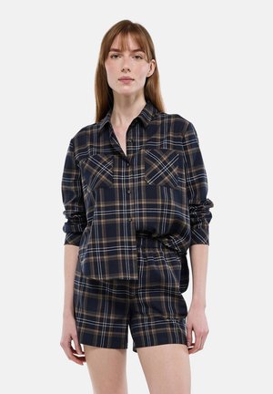 Femme portant une chemise à carreaux bleu marine et marron à manches retroussées, associée à un short à taille élastique assorti, debout devant un fond uni.