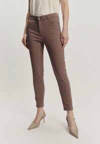 Brązowe dopasowane spodnie wykonane z elastycznego materiału, z wysokim stanem, wąskimi nogawkami i pięcioma kieszeniami. Noszone z beżowymi szpilkami.