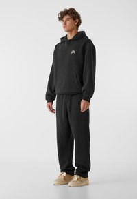 Crna dukserica s kapuljačom i kengur džepom, s malim ru�žičastim logotipom. Uparena s odgovarajućim crnim trenirkama i bež tenisicama s teksturiranim detaljima.