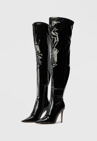Bottes en cuir verni noir au-dessus du genou avec un bout pointu et un talon haut élancé. Dotées d'une finition brillante et d'une fermeture éclair sur le côté extérieur.