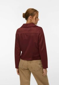Femme aux cheveux auburn attachés en chignon, portant une veste bordeaux et un jean beige, vue de dos sur un fond blanc uni.