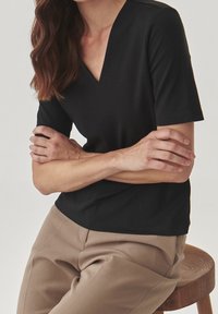 Schwarzes Kurzarm-Oberteil mit V-Ausschnitt aus glattem Stoff, kombiniert mit beigefarbenen, maßgeschneiderten Hosen, sitzend auf einem Holzstuhl.