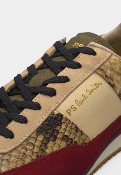 Κοντινό πλάνο αθλητικού παπουτσιού με μαύρα κορδόνια, μπεζ καστόρι, σχέδιο δέρματος φιδιού, κόκκινη καστόρι περιμετρικά και χρυσό λογότυπο "PS Paul Smith" στην πλαϊνή επιφάνεια.