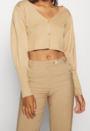 Femme portant un pull court beige boutonné à manches longues et un pantalon beige taille haute avec une fermeture par bouton.