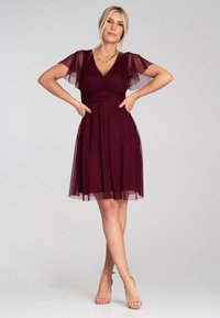 Burgunderfarbenes Kleid mit transparenten, flatternden Ärmeln, V-Ausschnitt und tailliertem Oberteil. Der fließende Rock reicht bis über das Knie. Schlichte Accessoires ergänzen das Outfit.