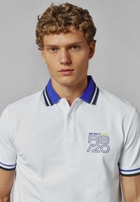 Balta polo marškinėliai su mėlynos ir pilkos spalvos juostelėmis ant apykaklės ir akcentinėmis rankovėmis. Priekinis logotipas rodo "RED BULL RACING RB/20" tamsiai mėlyna ir geltona spalvomis.