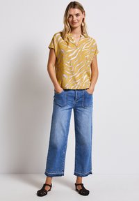 Top giallo a maniche corte con strisce bianche a zebra, abbinato a jeans a gamba larga azzurri con tasche e orlo grezzo. Sandali neri.