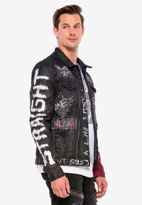 Zwarte denim jas met witte en rode grafische tekst. Bevat patches, studs en een verouderd ontwerp. Rits aan de voorkant en knoopzakken.