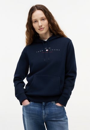 Mladá žena oblečená v námorníckej modrej mikine Tommy Jeans s nápisom New York City a modrými džínsami, stojaca s rukami vreckách na bielom pozadí.