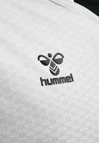 Czarne logo Hummel ze stylizowaną pszczołą i tekstem na białym, fakturowanym tle z tkaniny.