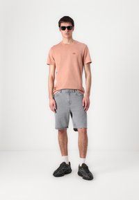 Lyserød bomull t-skjorte med rund hals og liten logo, kombinert med grå denimshorts og svarte joggesko, mot en ensfarget bakgrunn.