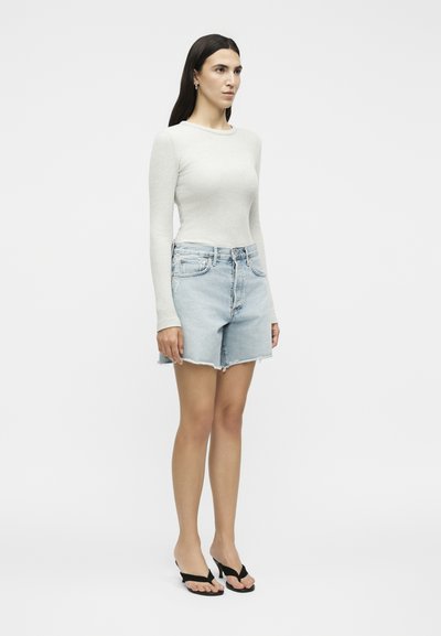 Γυναίκα στέκεται προς τα εμπρός, φοράει λευκή μπλούζα με μακριά μανίκια, ανοιχτό μπλε τζιν σορτς με ξεφτισμένα τελειώματα και μαύρα πέδιλα με τακούνι.