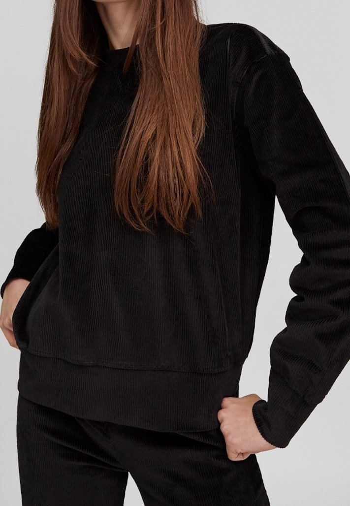 Femme portant un haut à manches longues en velours côtelé noir et un pantalon assorti en velours côtelé noir, une main sur la hanche et de longs cheveux bruns.