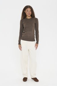 Femme aux cheveux bouclés se tient debout dans un pull marron à manches longues, un pantalon large blanc et des mocassins marron devant un fond blanc.