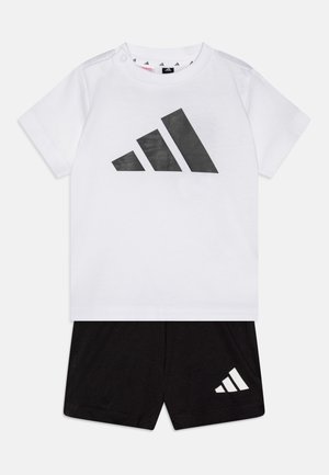 Baltas kokvilnas T-krekls ar lielu melnu Adidas logo, apvienots ar melniem šortiem, kuriem uz kājas ir neliels balts logo.