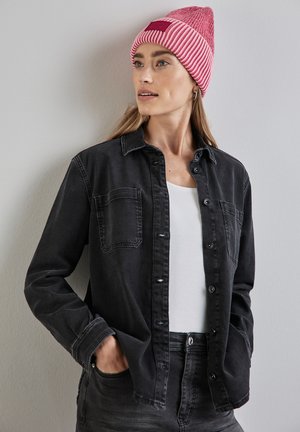 Schwarzes Denim-Hemd mit Knopfverschluss und zwei Brusttaschen, getragen über einem weißen Top. Begleitet von einer rosa gestreiften Strickmütze.