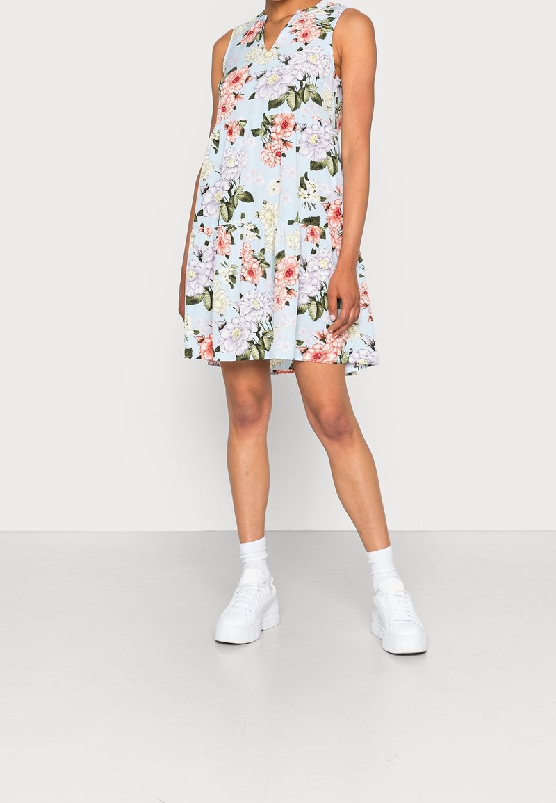 Femme portant une robe sans manches bleu clair à fleurs roses et blanches, des chaussettes blanches et des baskets blanches à plateforme, debout sur un fond uni.