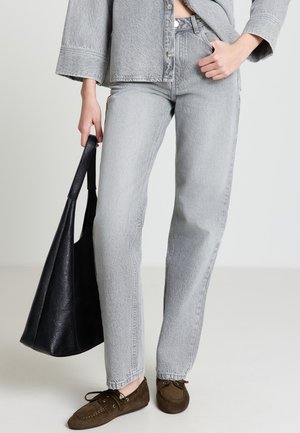 Person trägt hellgraue Jeans und Jacke, hält eine schwarze Ledertasche und trägt braune Wildleder-Schnürschuhe.