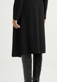 Schwarzes Kleid mit langen Ärmeln und einem leicht ausgestellten Saum, kombiniert mit glänzenden schwarzen Leggings mit Schlangenleder-Muster. Strukturierte Akzente an den Manschetten.