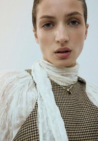 Licht stoffen sjaal met ruches valt rondom een geruit bruin textiel topje. Een gouden ketting met een rechthoekige hanger bij de hals.