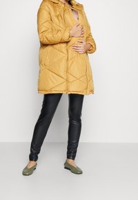 Gul vadderad jacka med huva, diamantmönstrad quiltning, dragkedja fram, kombinerad med svarta leggings och gröna platta skor.