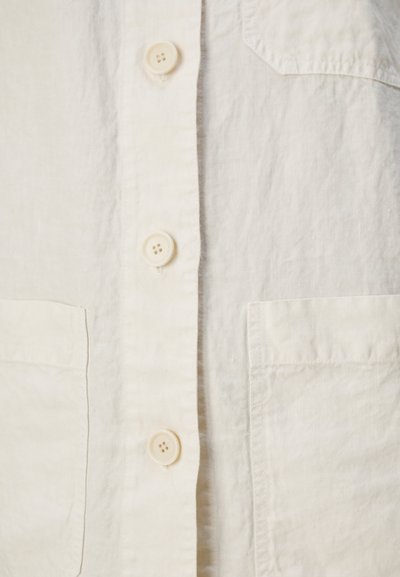 Chemise en lin beige clair avec un boutonnage à l'avant, deux poches poitrine et une texture légèrement froissée. Les boutons sont d'un crème clair avec quatre trous.