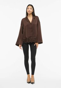 Brun sateng overdimensjonert skjorte med krage, knapper foran og en dekorativ blondekant, kombinert med svarte leggings og hæler.