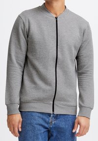 Veste zippée grise texturée avec un col rond, dotée d'une fermeture éclair noire et de poignets côtelés, associée à un jean en denim bleu.