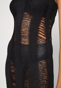 Dettaglio di un abito lavorato a maglia nera con strisce verticali a maglia aperta che rivelano la pelle e la biancheria intima nera sottostante.