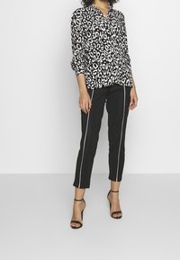 Bluse i sort og hvid leopardprint med krave, parret med sorte bukser med hvide side-striber og bøjler i sort.